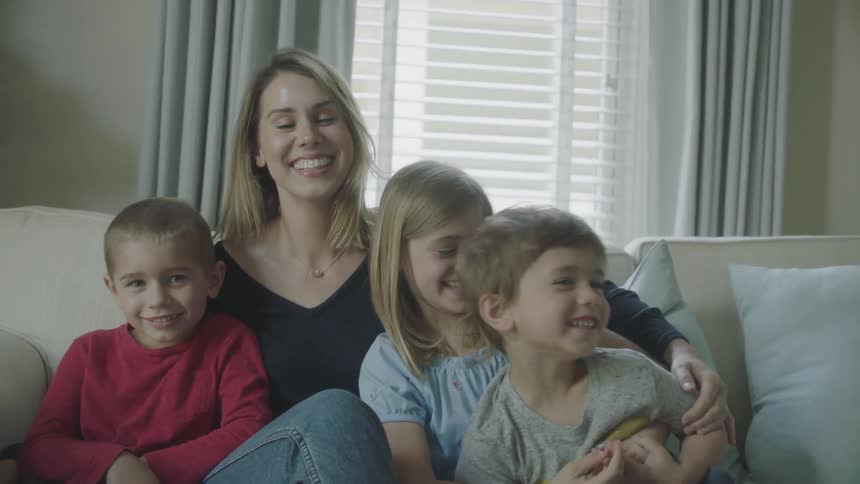 A mom sits on the sofa with her arms around her three children as they laugh together
