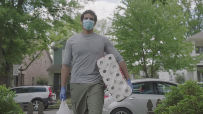 A man wearing a mask and gloves takes a grocery bag and toilet paper out of the trunk and carries them toward a house