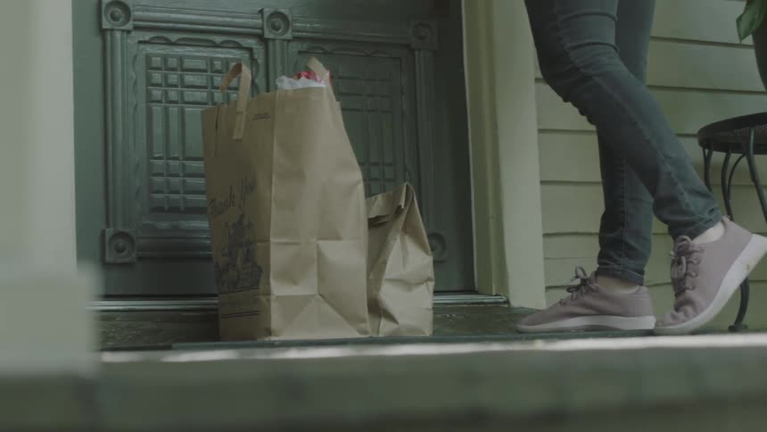 A woman wearing protective gloves leaves paper bags of groceries at the front door