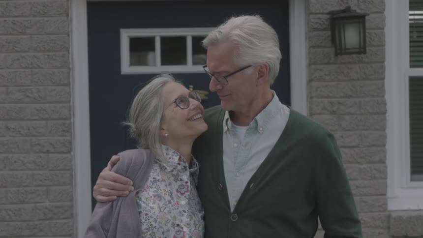 An elderly man has his arm around a woman and they look at one another and smile