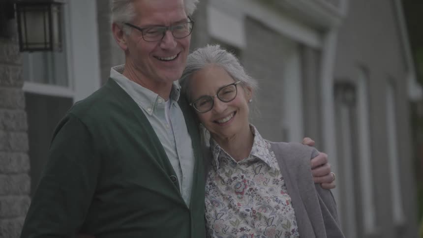Elderly man and woman stand and hold each other in front of the house