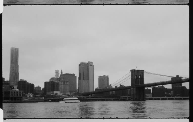 Old time footage of a body of water with a boat going under a bridge with a large city in the background