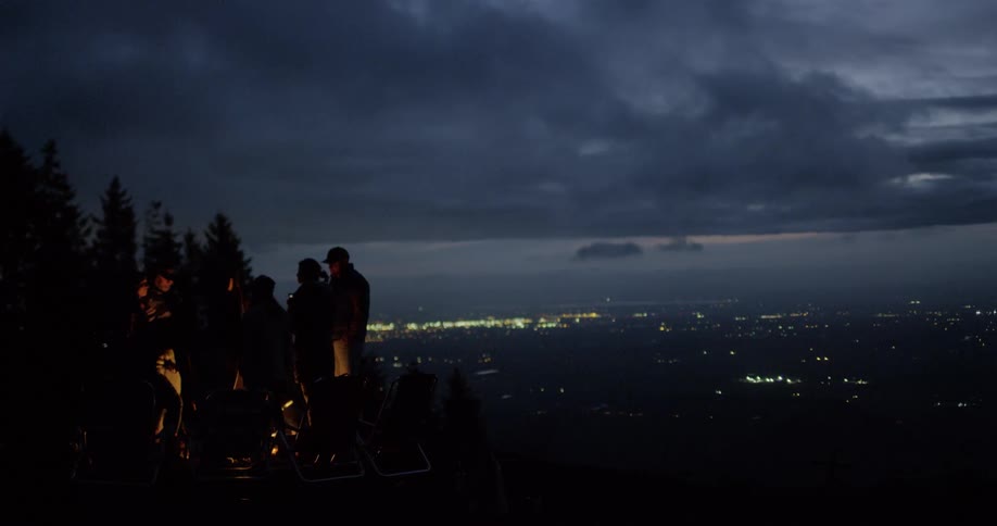 A small group of people stand around a fire with the city skyline in the background