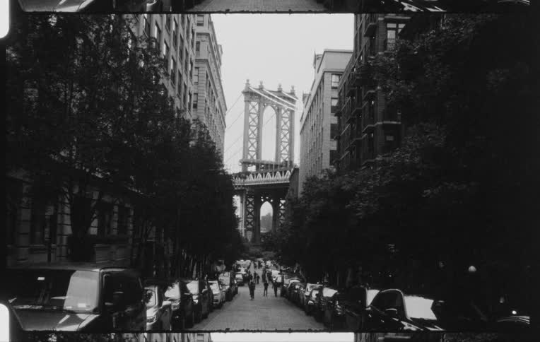 Pedestrians move among cars parked at side of road with suspension bridge at end of street