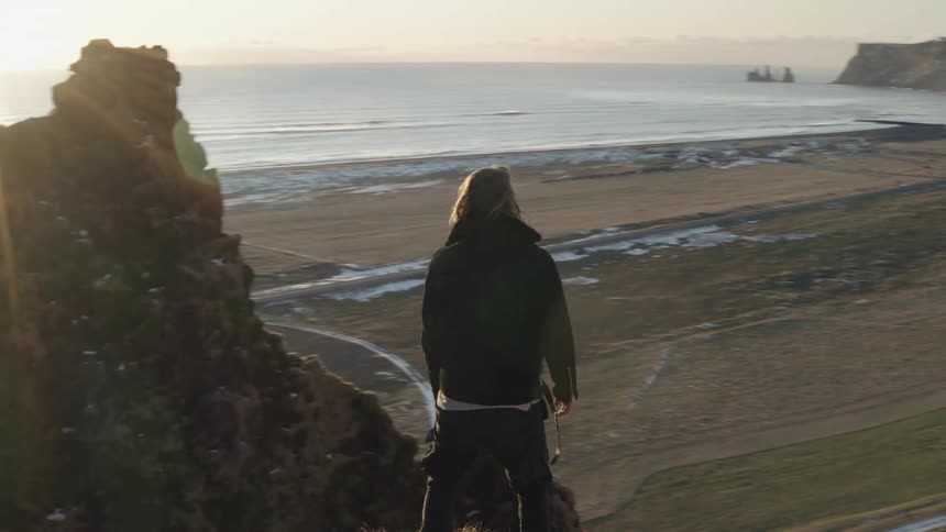A man stands on top of a high mountain cliff oceanside
