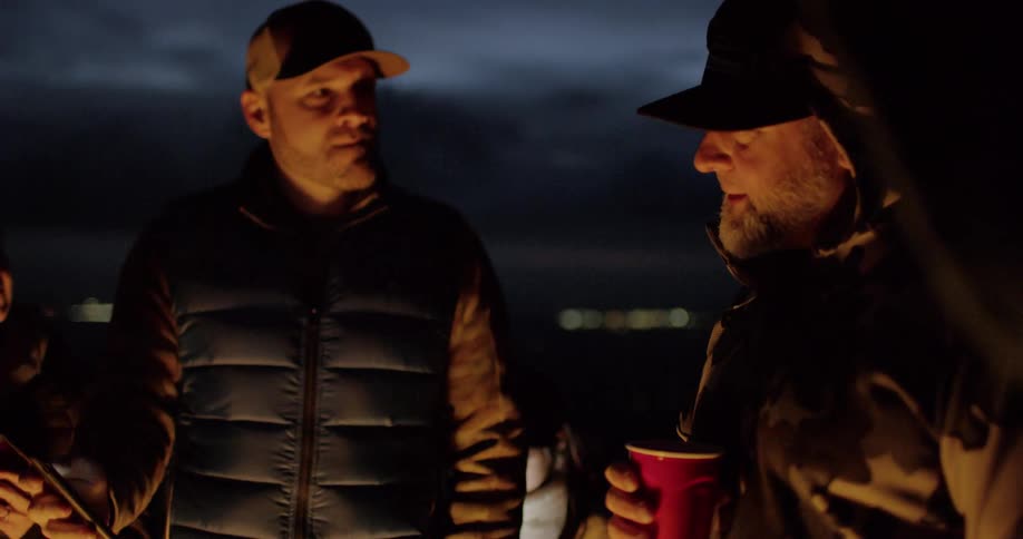 A man shows his friend something on his phone as they stand talking with firelight shining and glowing