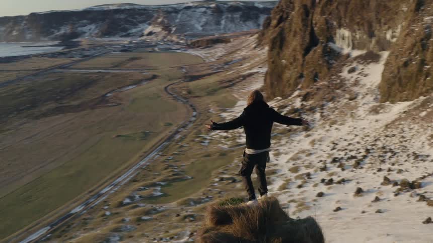 Man stands on peak with arms outstretched staring out to sea near sand covered mountain slope