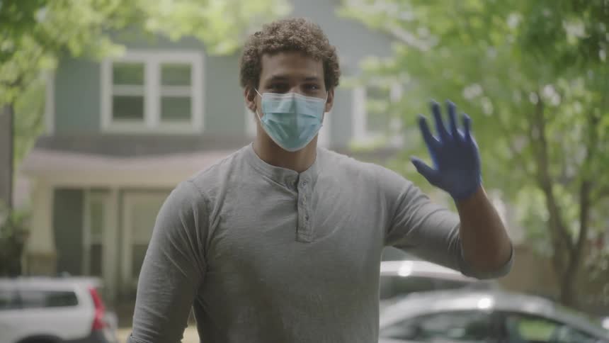 Young man wearing a face mask delivering a paper bag on front porch and waving