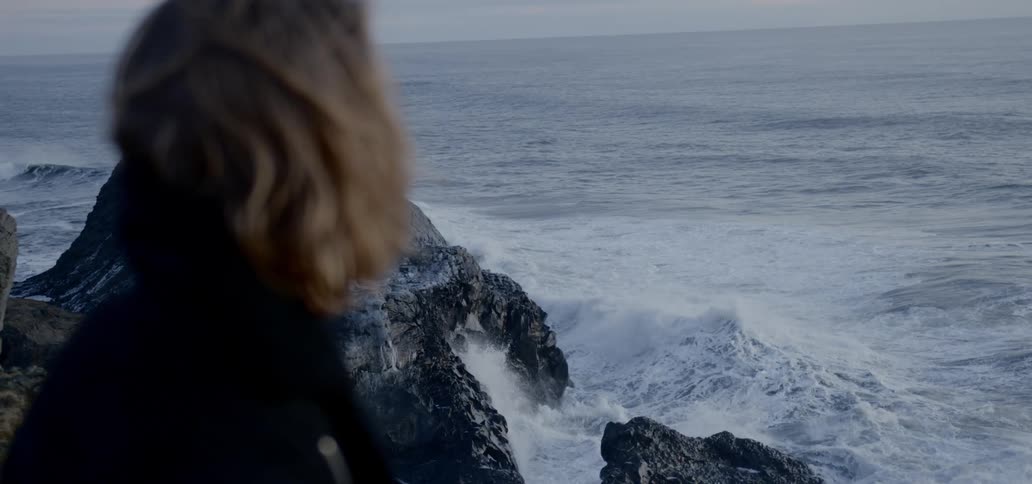 A woman sits on a cliff overlooking a rough sea