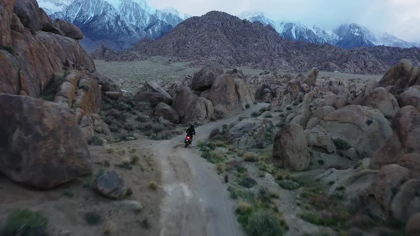 Motorcyclist rides along narrow dirt road among rough terrain strewn with boulders near mountain ranges