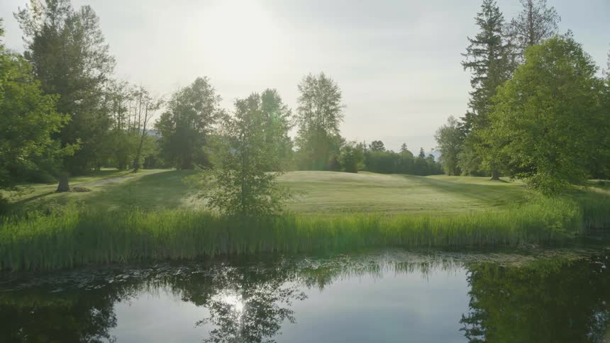 Drone footage flying over a still pond toward a wooded area with cut grass and rolling hills