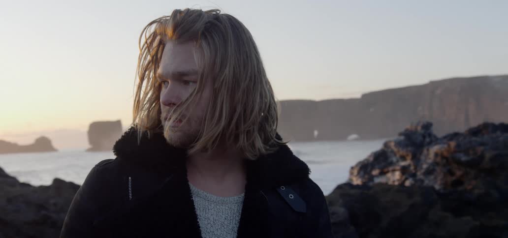 Blonde long haired man standing on the rocky coast of a beach at sunset