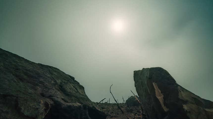 Sun in the sky and clouds pass over rocks and tree branches in a barren area
