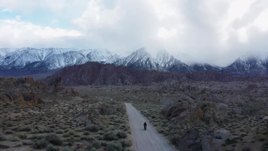 A person drives a motorcycle towards snow capped mountains