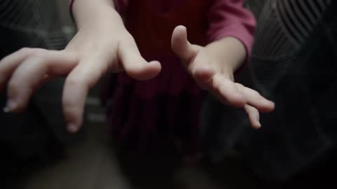 A young white child stands indoors while stretching her hands in front of her