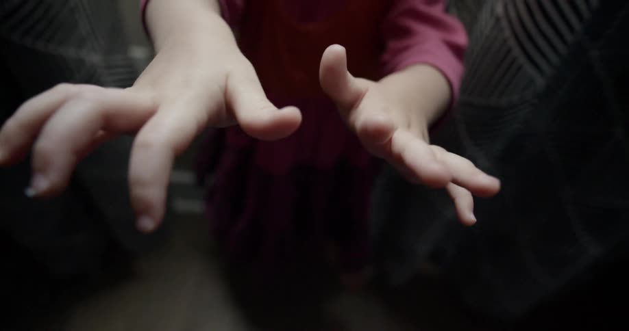 A young white child stands indoors while stretching her hands in front of her