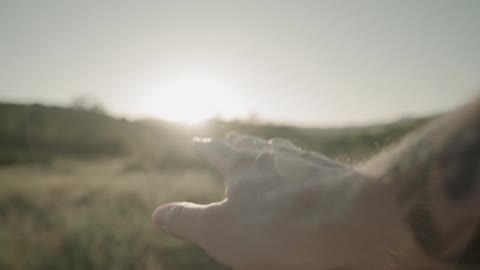 A mans hand outdoors with a grassy field and the sun in the background