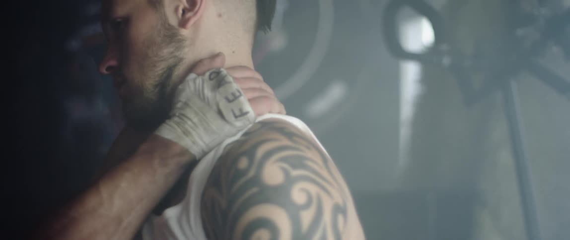 A white male with a tattoo rubs his shoulder and neck as he sits in a gym