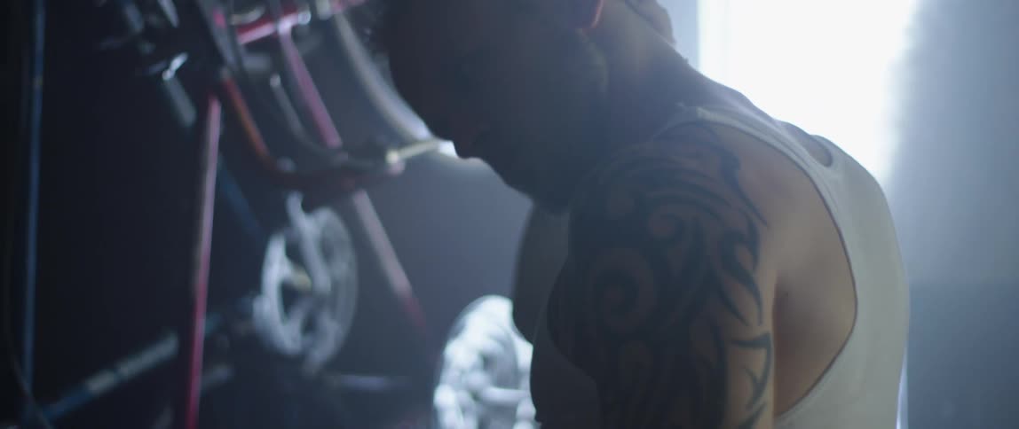 A man with a tattoo on his shoulder and arms rubs the back of his neck and head while standing in a garage