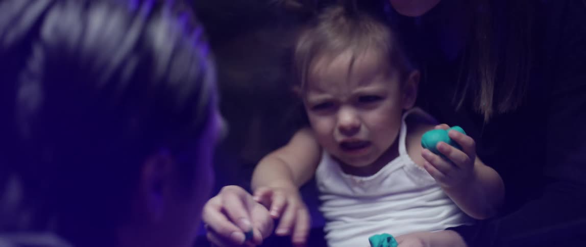 A man taps on a glass barrier to make his daughter smile as she sits with her mom playing