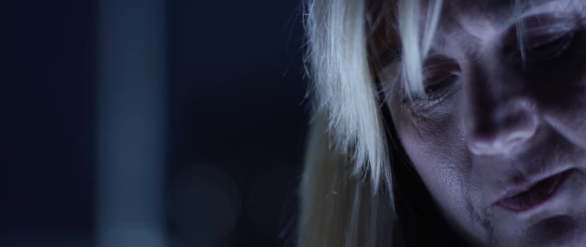 A woman looks down and mumbles in a room with a blue light