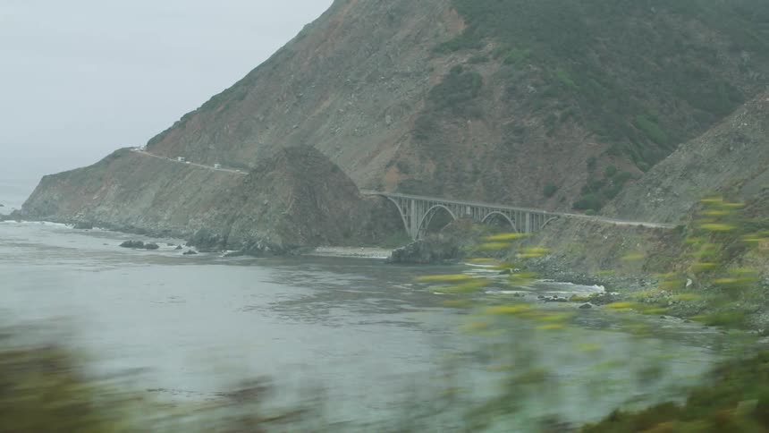 Vehicles drive along road hugging mountain as waves crash on shore of foggy day