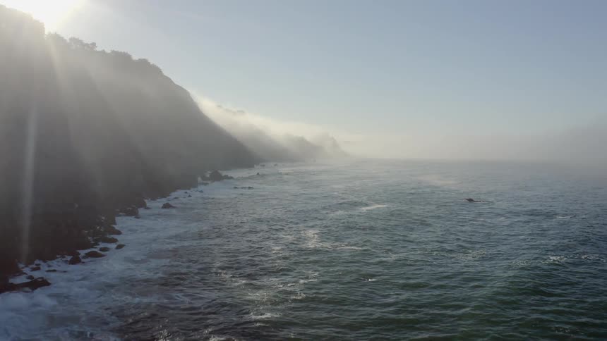 Sunshine glares through fog as waves crash along base of cliff
