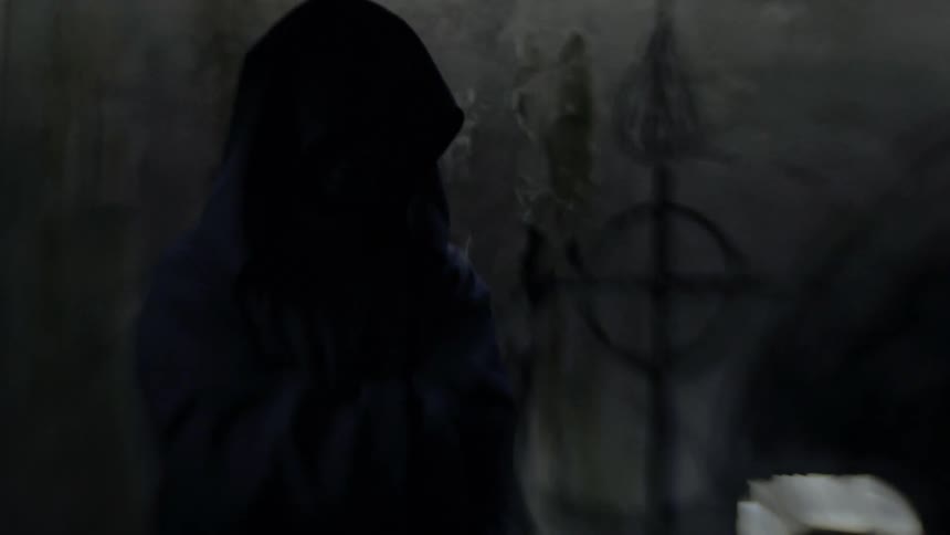 A dark figure wearing a hood stands in a dark chamber