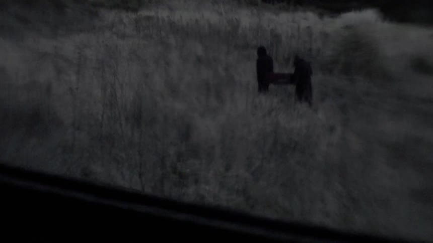 Two robed figures carry a coffin through the grass viewed from a window
