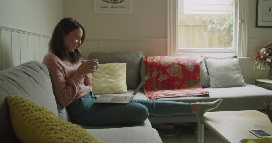 A woman sitting on the couch holding a cup of tea talks with someone using her laptop