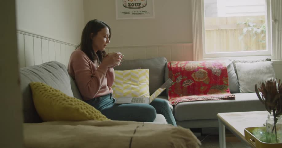 Woman sitting in couch in the living room uses laptop while drinking tea