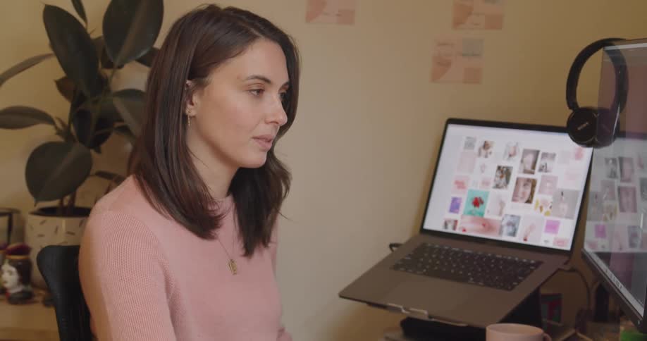 A white woman sits at a desk with a computer and writes with a pen