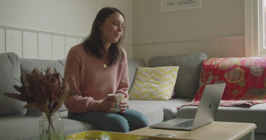 Woman sits on couch drinking from a coffee mug as she talks via her laptop