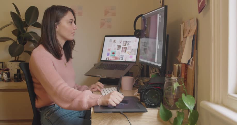A white woman sits at a desk indoors working on a computer with a writing pad