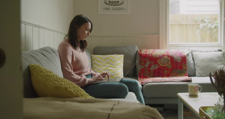 A woman sits on a home couch and works on a laptop