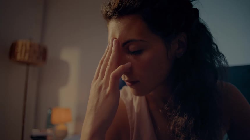 A woman sitting on the edge of a bed leaning forward and rubbing her forehead with eyes closed