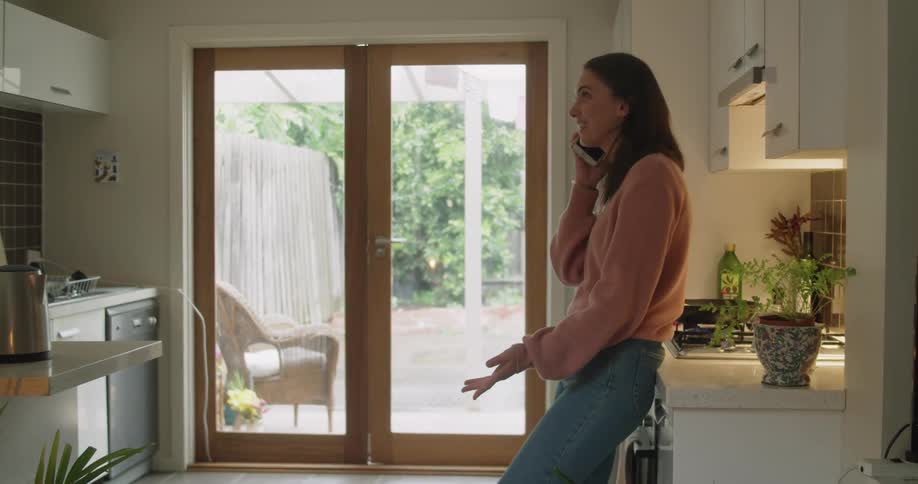 A woman laughs while talking on her smart phone while standing in the kitchen