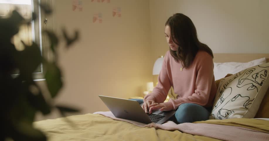 Woman sitting on the bed types on a laptop in her room