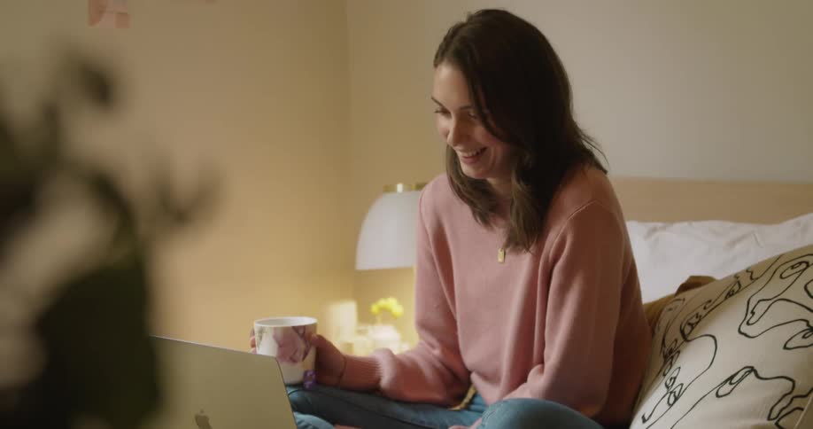 Woman sitting on bed holding a mug laughing watching a movie on laptop