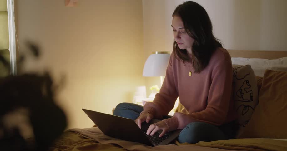Woman sits on a bed as she types on a laptop