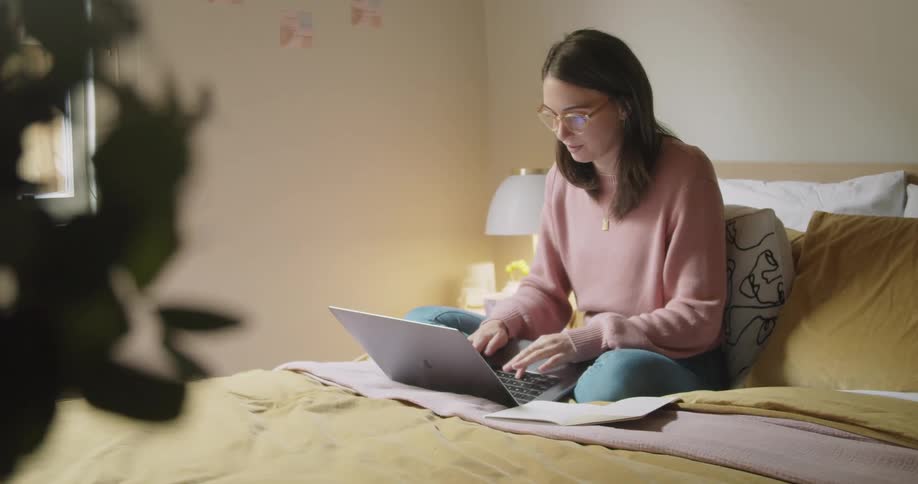 A woman sits and types on a laptop while sitting in bed