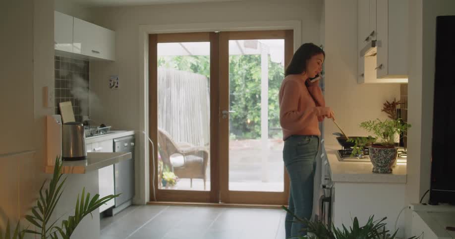 A woman in a kitchen talking on a smart phone while cooking