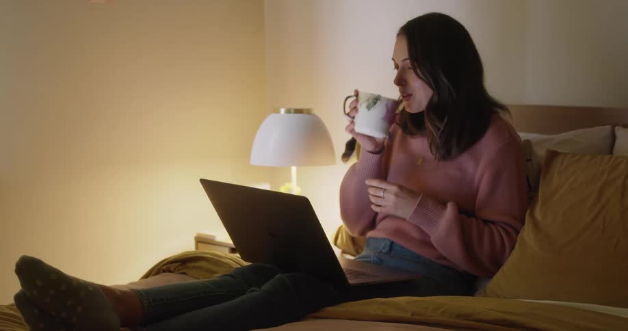 Woman drinking coffee watching a movie on laptop while relaxing on bed
