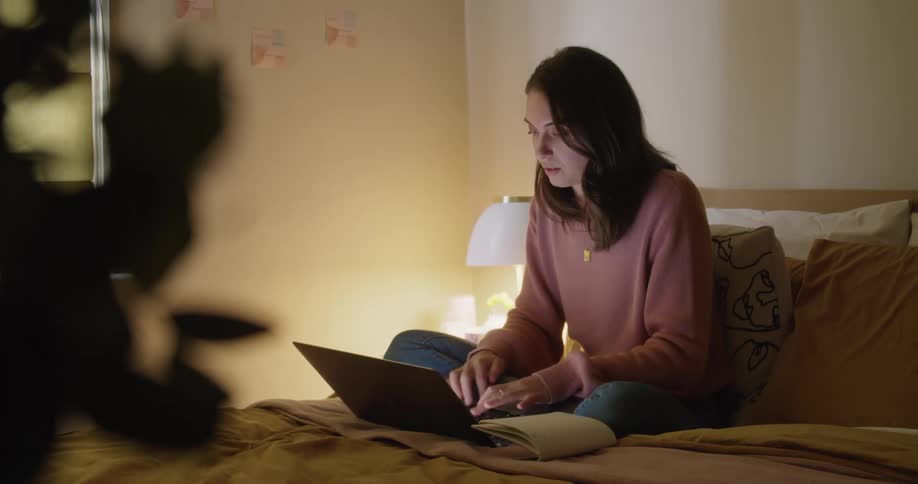 A woman sitting on a bed typing on a laptop