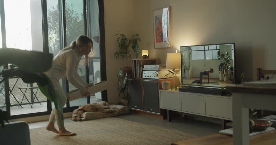 A woman unrolls a mat as she prepares to do yoga with an instructor on the television