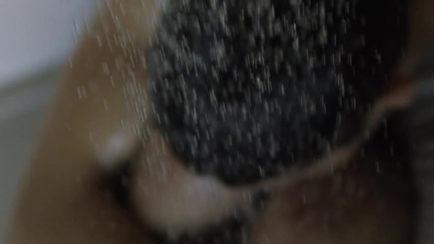 A person sits with arms wrapped around themselves as shower water falls on them