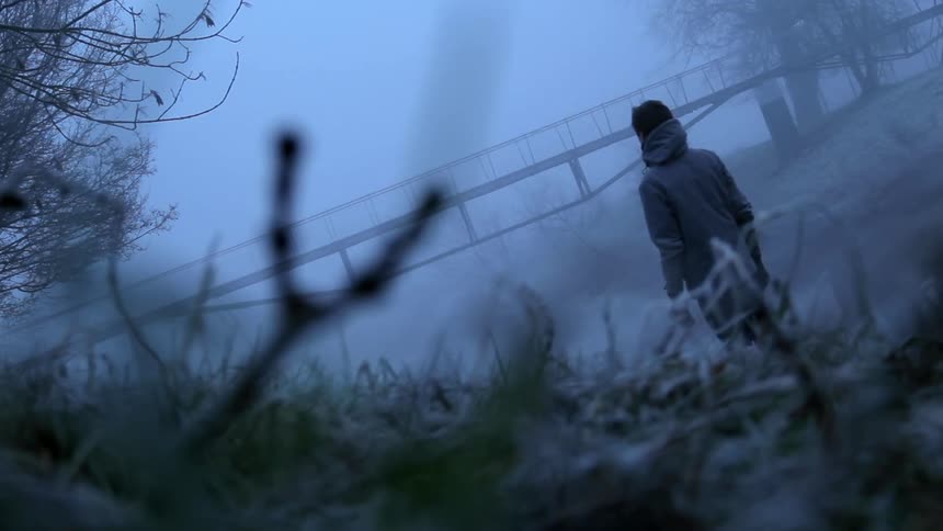 A person in a hoodie walks near a bridge in an eerie foggy forest
