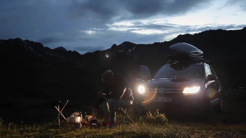 A man cooks on a camping stove while sitting in front of his suv which has its light on