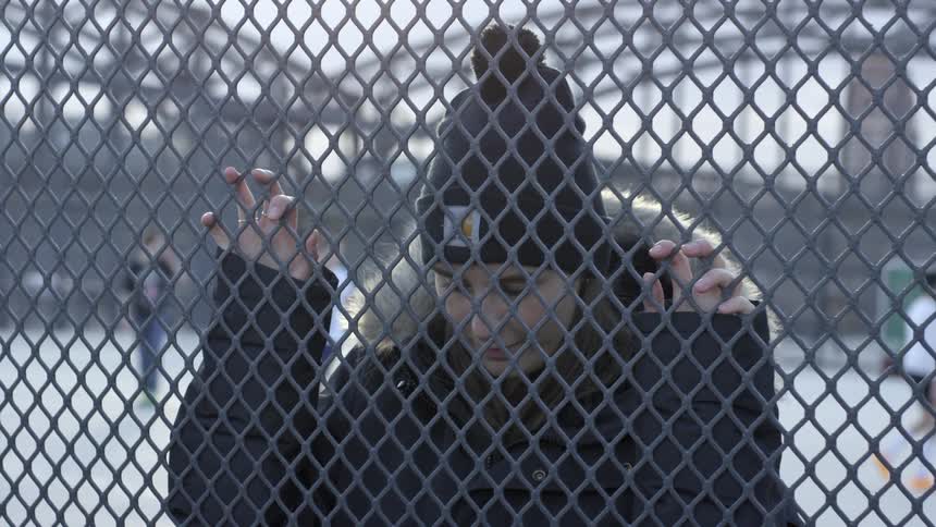 A white woman grabs a chain link fence and drops her head