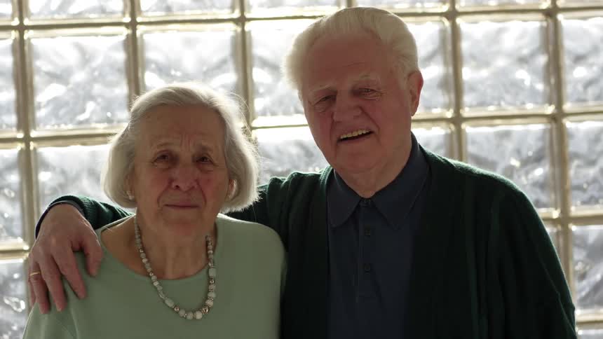 Elderly man and woman smile happily and pose for the camera in front of a frosted glass